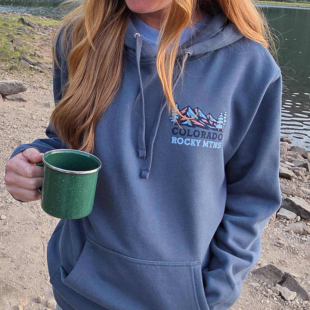 Person wearing a blue hoodie with 'Colorado Rocky Mtns' text and holding a green mug by a body of water.