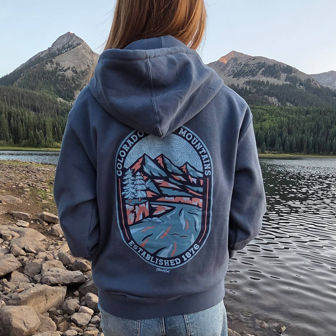 Person wearing a blue hoodie with a mountain design by a lake and mountains