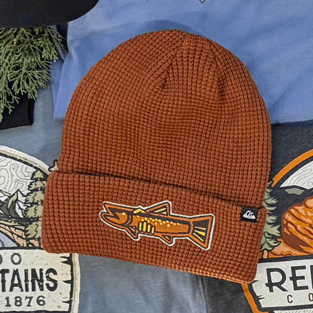 Rust colored beanie with fish patch on front displayed on top of other products.