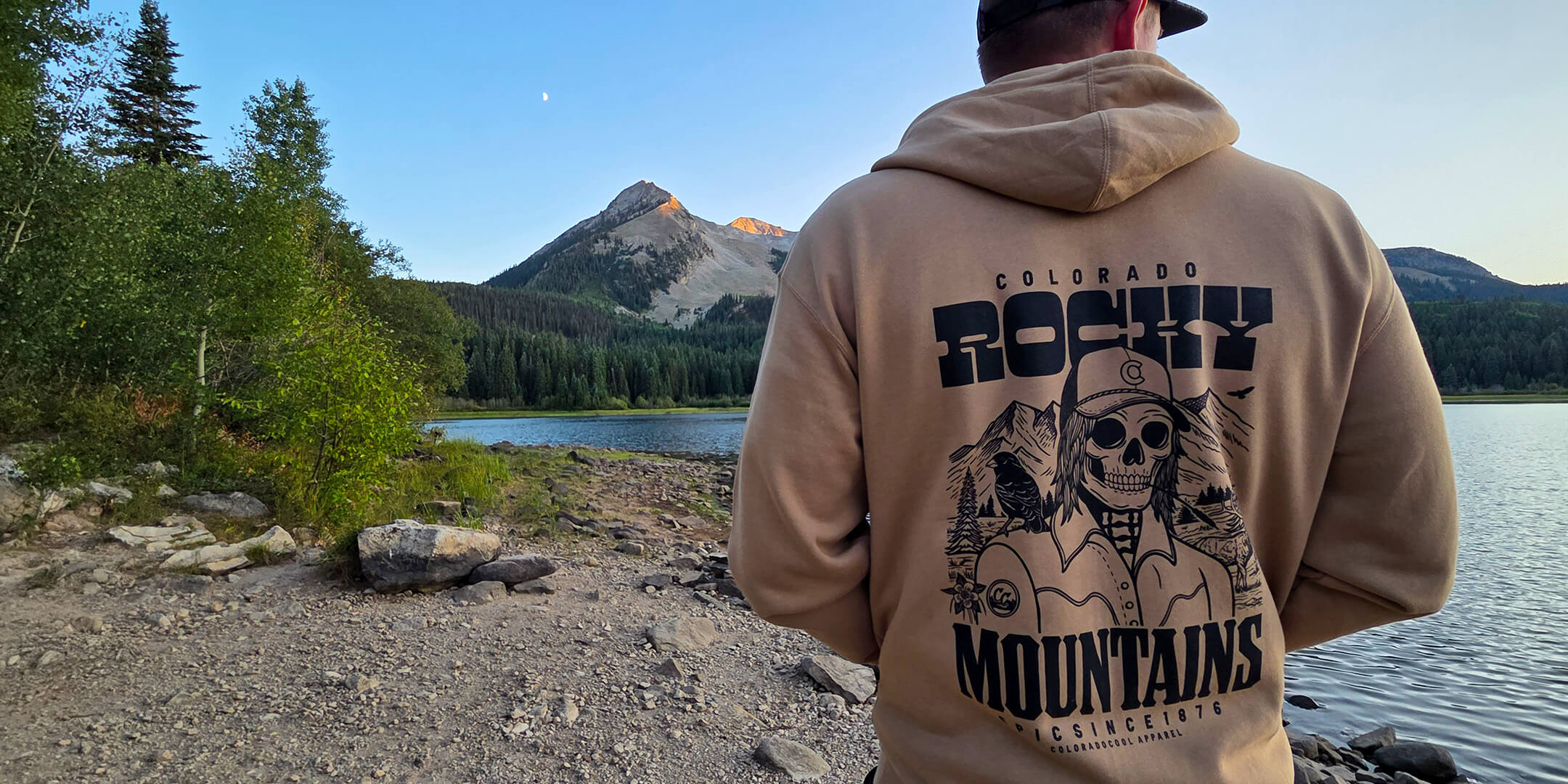 Person wearing a hoodie with 'Colorado Rocky Mountains' design by a lake and mountains.