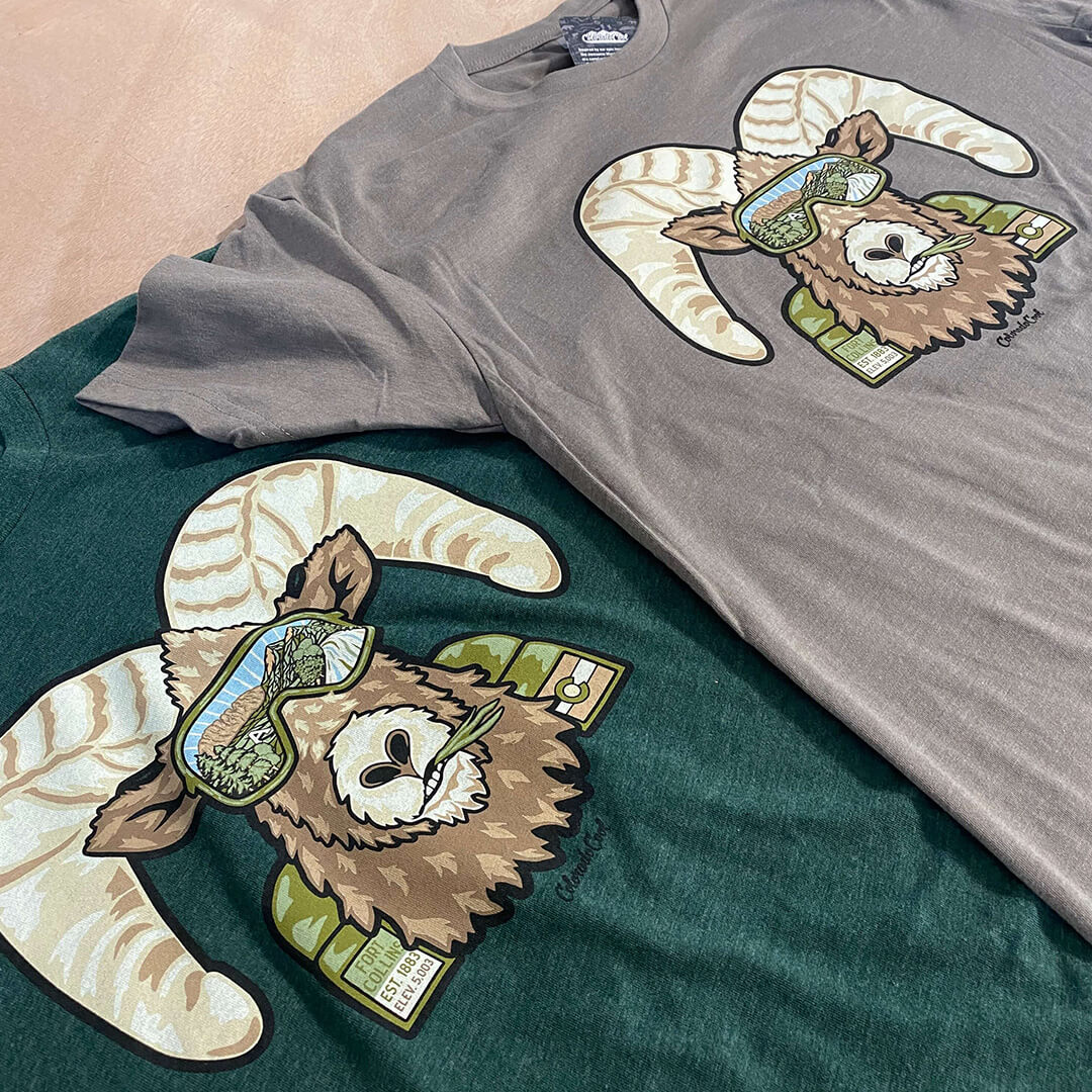 Image of two t-shirts laying flat on a table with a bighorn sheep (ram) on the front with views of Fort Collins in the ram's sunglasses